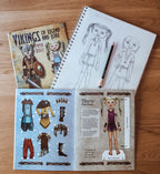 A children's craft activity book titled 'Vikings of Legend and Lore' shown with a spiral sketchbook, pencil, and an open page featuring a character sheet and outfit cutouts, all arranged on a wooden surface.