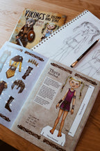 A children's craft activity book titled 'Vikings of Legend and Lore' shown open on a wooden table, featuring character sheets for 'Thyra the Dancerod' with illustrations of clothing and accessories, alongside a pencil and another sketchbook.