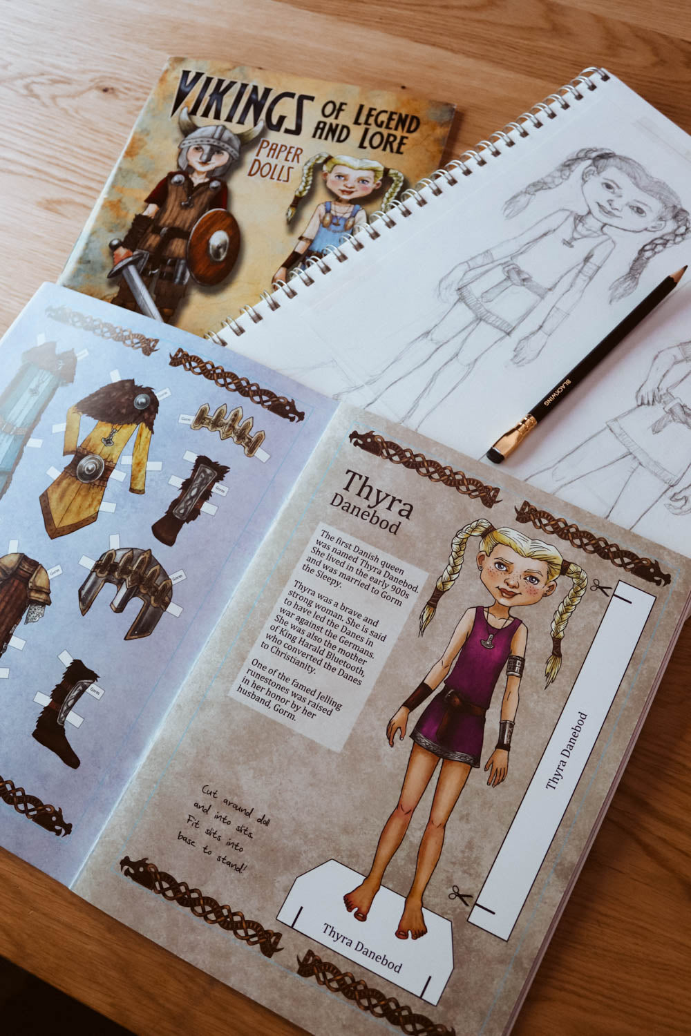 A children's craft activity book titled 'Vikings of Legend and Lore' shown open on a wooden table, featuring character sheets for 'Thyra the Dancerod' with illustrations of clothing and accessories, alongside a pencil and another sketchbook.