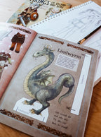 A close-up of an open craft or activity book showing a printed Lindwyrm dragon illustration and instructions, with a spiral notebook and pencil on a wooden surface.