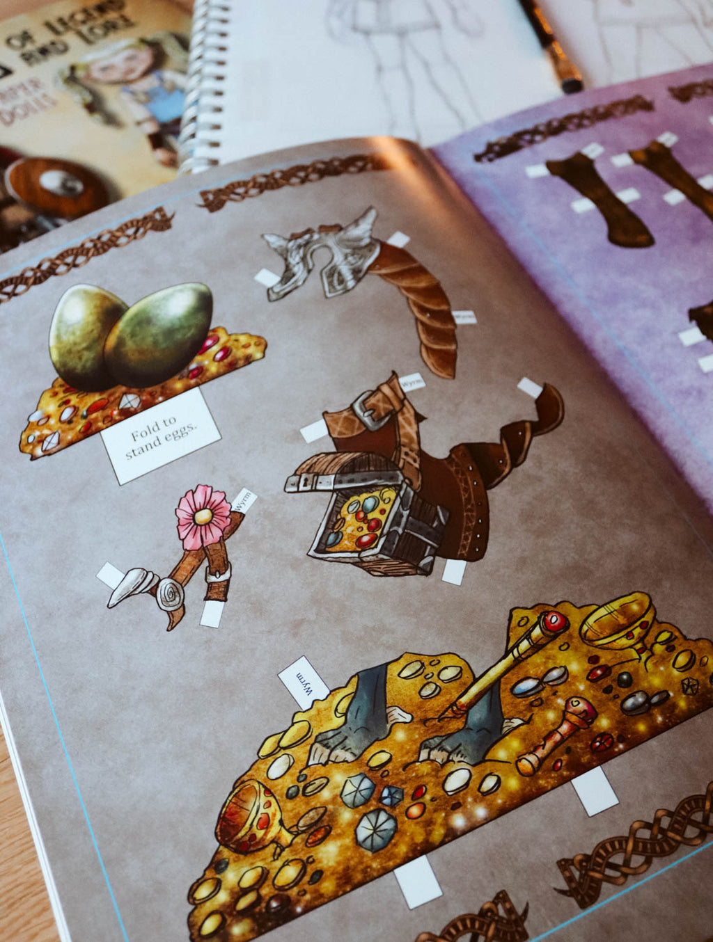 An open spiral-bound craft or activity book showing illustrated puzzle pieces, including a treasure chest, coins, a flower, and a small sign reading 'TINY TOAD STAMENTS,' photographed on a tabletop.