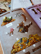 An open spiral-bound craft or activity book showing illustrated puzzle pieces, including a treasure chest, coins, a flower, and a small sign reading 'TINY TOAD STAMENTS,' photographed on a tabletop.