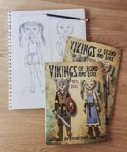 Two illustrated books titled 'Vikings of Legend and Lore' for paper dolls are arranged on a wooden surface, with a pencil on top and a sketchbook showing a pencil drawing of one of the characters.