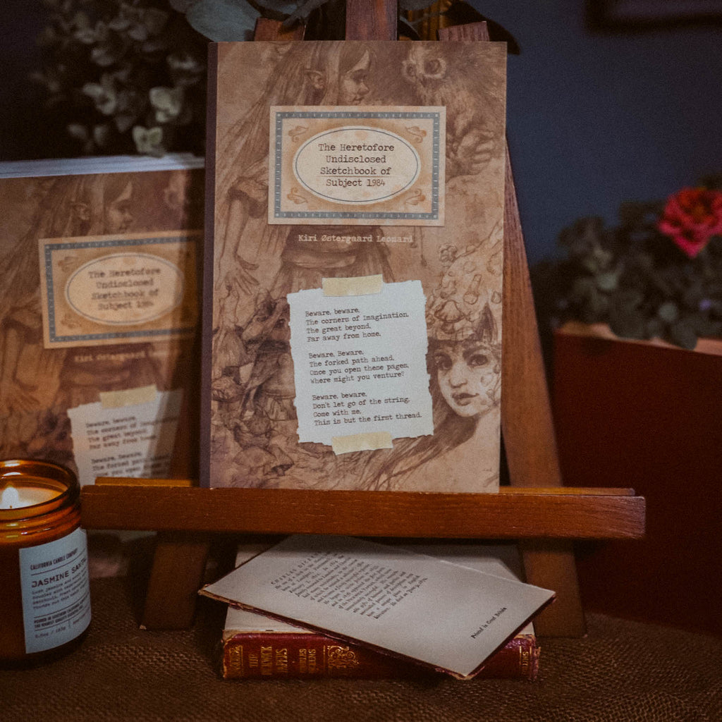 A vintage-style illustrated card or booklet titled 'The Infatuated Misunderstanding Sketchbook of Robert Kin' displayed on a wooden stand, with a matching copy in the background and a small candle nearby on a table.