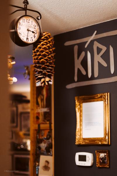 A dark wall decorated with a large pinecone hanging from a black wall clock, a gold-framed picture, and the word 'KIRI' written in white on a black background.