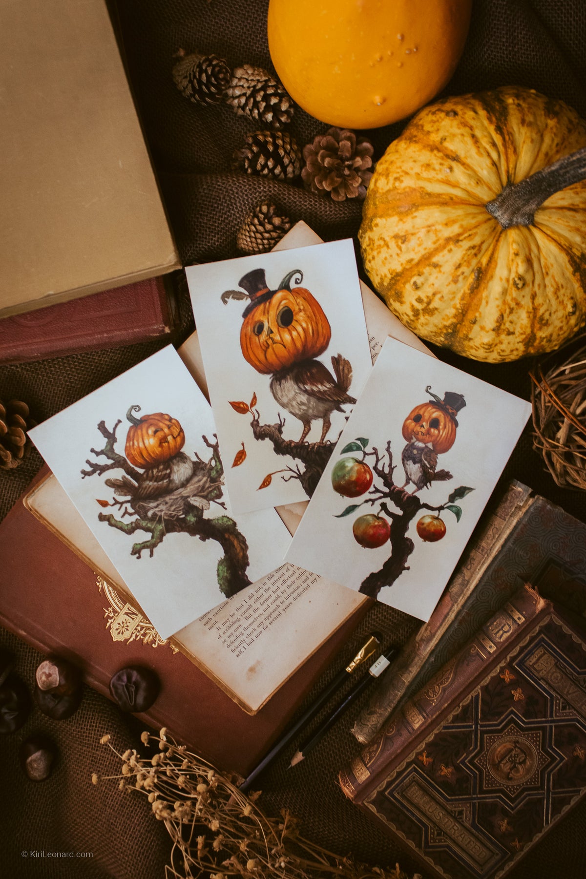 Three art prints featuring whimsical illustrations of pumpkins and birds on autumn trees, styled in a flat lay with vintage books, pinecones, dried grass, and pumpkins.