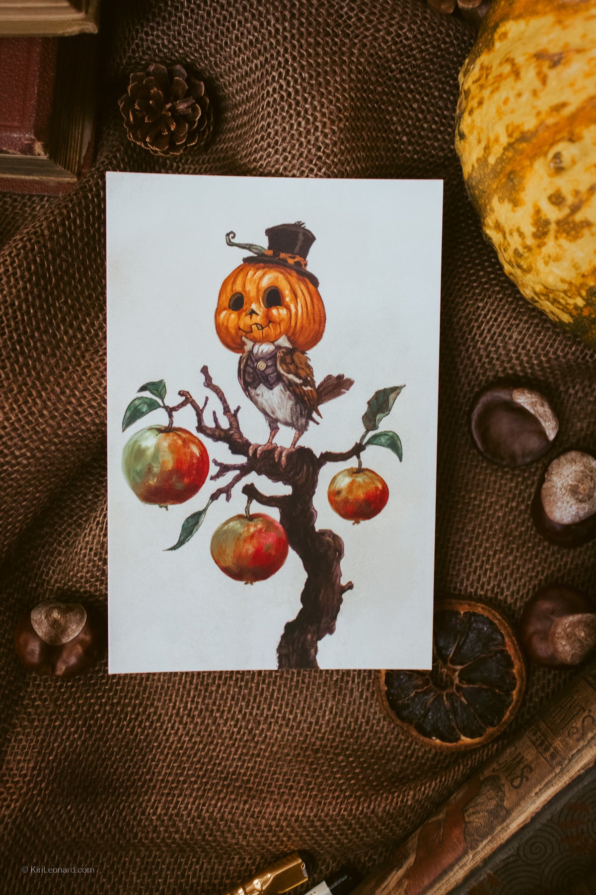 A square art print featuring a watercolor illustration of a bird with a carved pumpkin head and small top hat perched on an apple tree branch, styled on a brown burlap surface with autumn props.