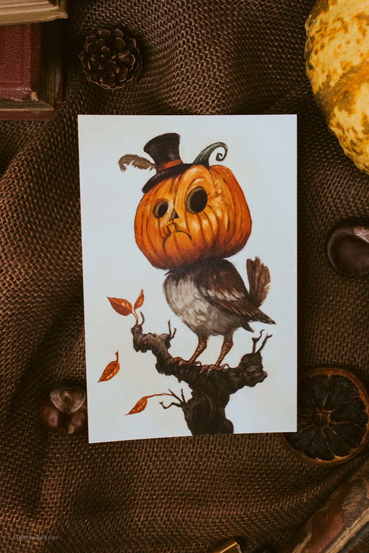A vertical art print featuring a whimsical illustration of a small brown bird with a carved orange pumpkin head wearing a tiny black top hat, perched on a branch with scattered autumn leaves. The print is displayed flat on a textured brown burlap fabric surrounded by seasonal props like a pinecone, dried orange slice, acorns, and a yellow gourd.