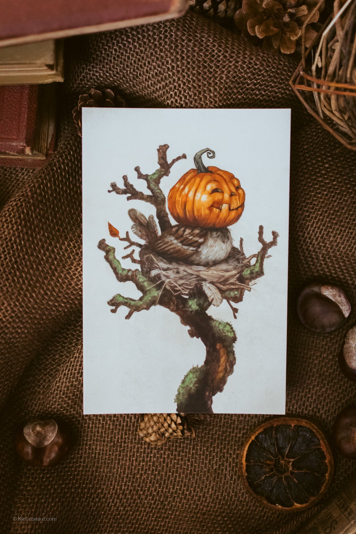 A vertical art print featuring a carved orange pumpkin on a gnarled tree branch with a bird's nest, displayed flat on a textured brown burlap surface with autumn props.
