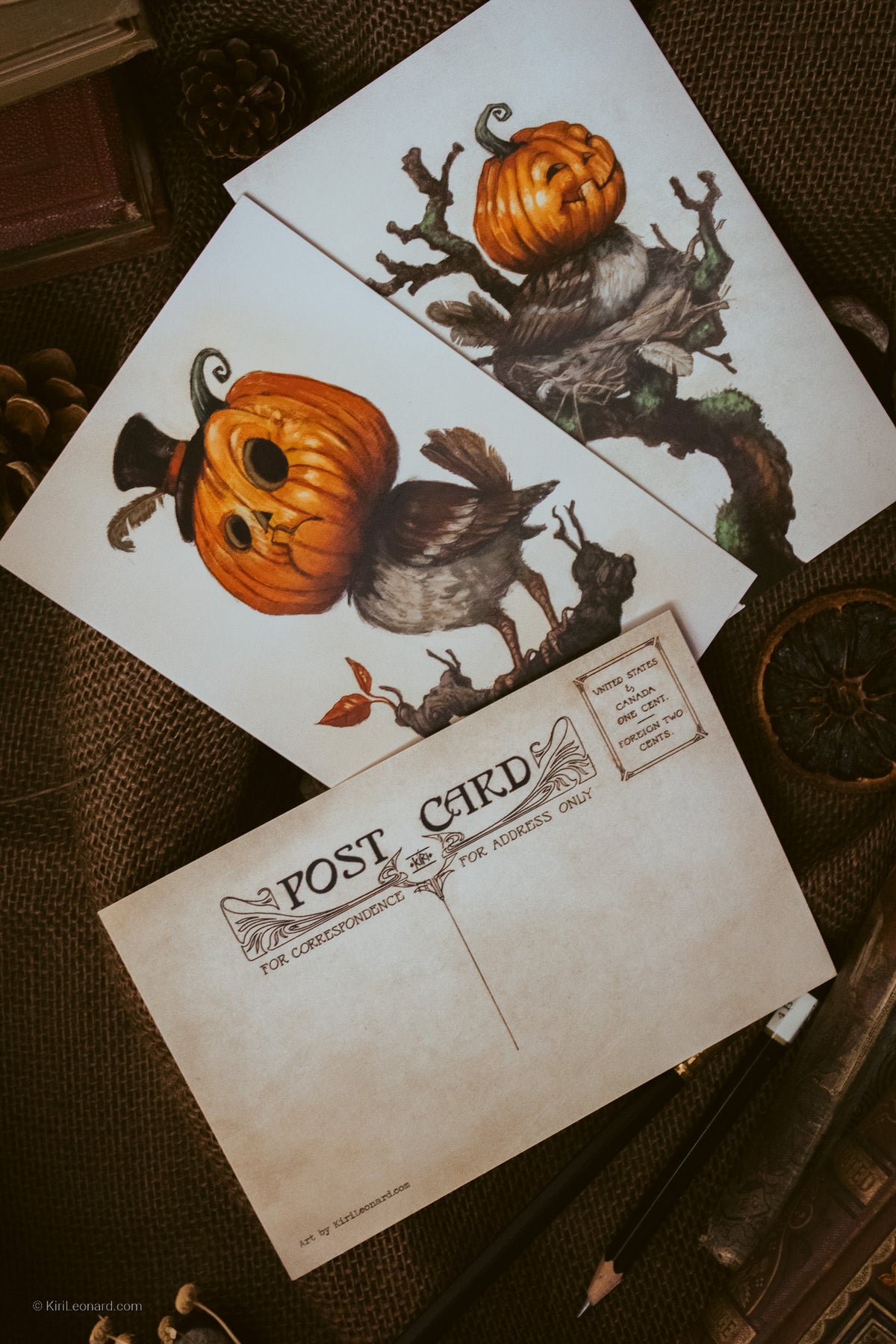 Two vintage-style Halloween postcards with illustrated jack-o'-lantern pumpkins and a bird, arranged on a textured brown surface with a blank postcard in front and autumn props around them.