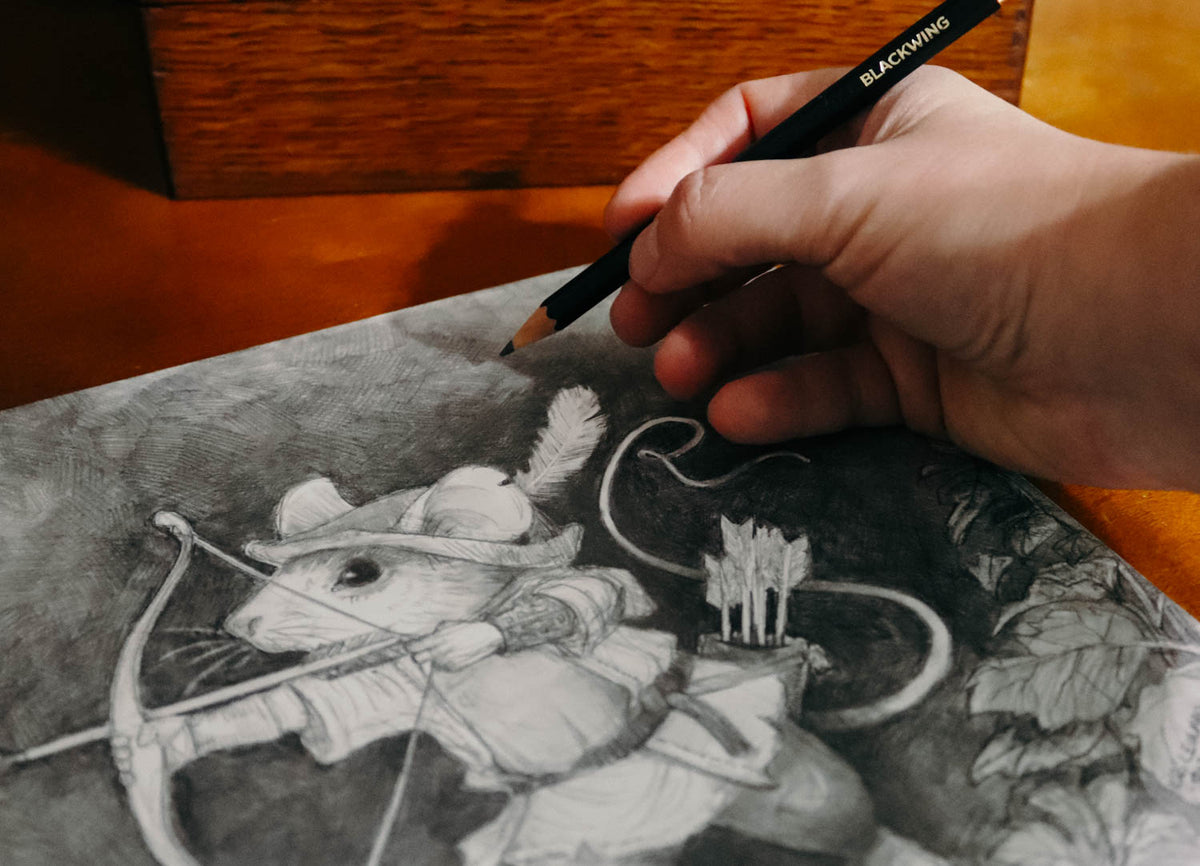 A close-up of a hand holding a black pencil with the brand name 'BLACKCROW' visible, drawing a detailed black and white sketch of a mouse archer on paper over a wooden surface.