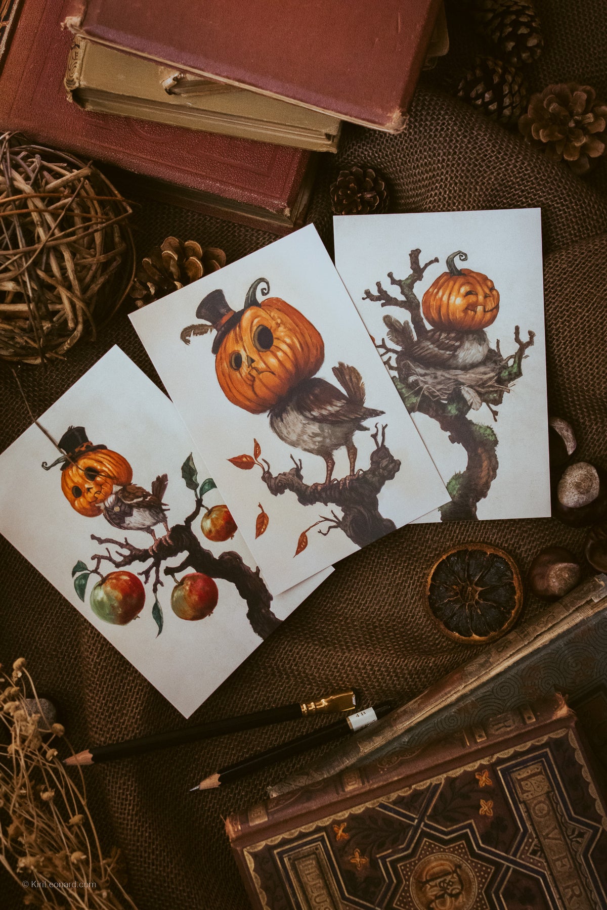 Three art prints featuring whimsical illustrations of pumpkins on tree branches with birds and apples, arranged in a styled flat-lay with vintage books, pencils, and dried botanicals on a brown burlap background.