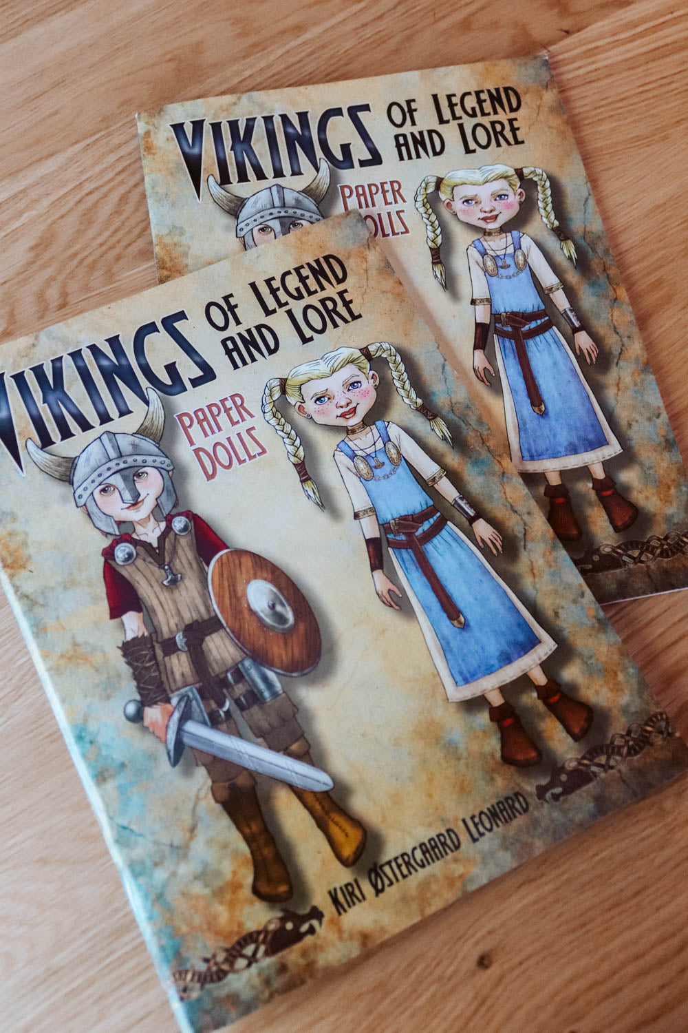 Two copies of the book 'Vikings of Legend and Lore: Paper Dolls' by Kiri Oestergaard Leonard, featuring illustrated Viking characters on the cover, displayed on a light wooden surface.