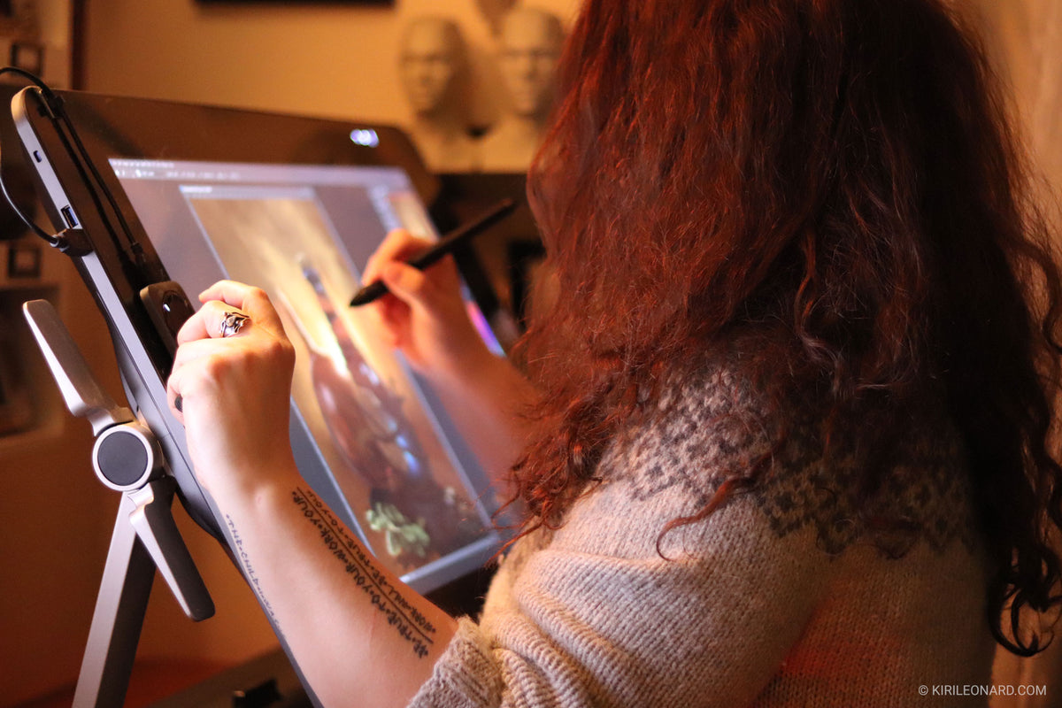 A person with long curly brown hair is shown from behind, using a stylus on a digital drawing tablet in a warm indoor setting.