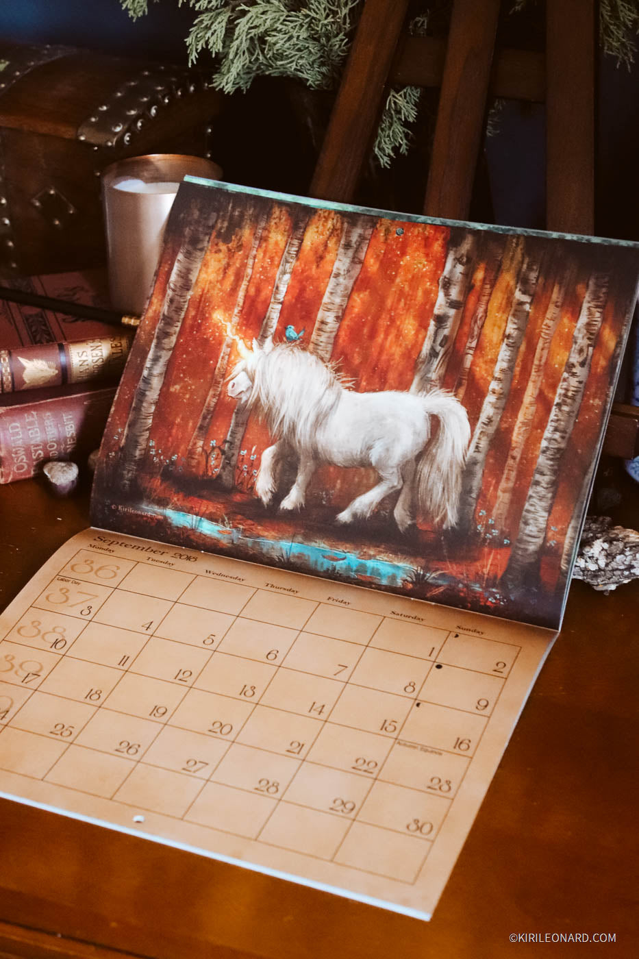 An open wall calendar or desk calendar on a wooden table, showing a large printed illustration of a white horse in front of a fiery orange and brown forest scene. The lower portion of the calendar page shows numbered days in a grid.