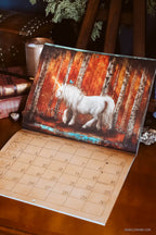 An open wall calendar or desk calendar on a wooden table, showing a large printed illustration of a white horse in front of a fiery orange and brown forest scene. The lower portion of the calendar page shows numbered days in a grid.