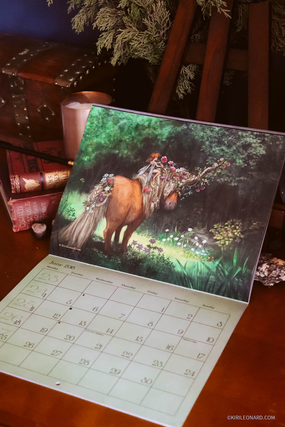 A desk calendar displayed on a wooden table, featuring a colorful illustration of a pony in a forest on the cover page. The calendar pages are fanned out below, showing the monthly grid layout.