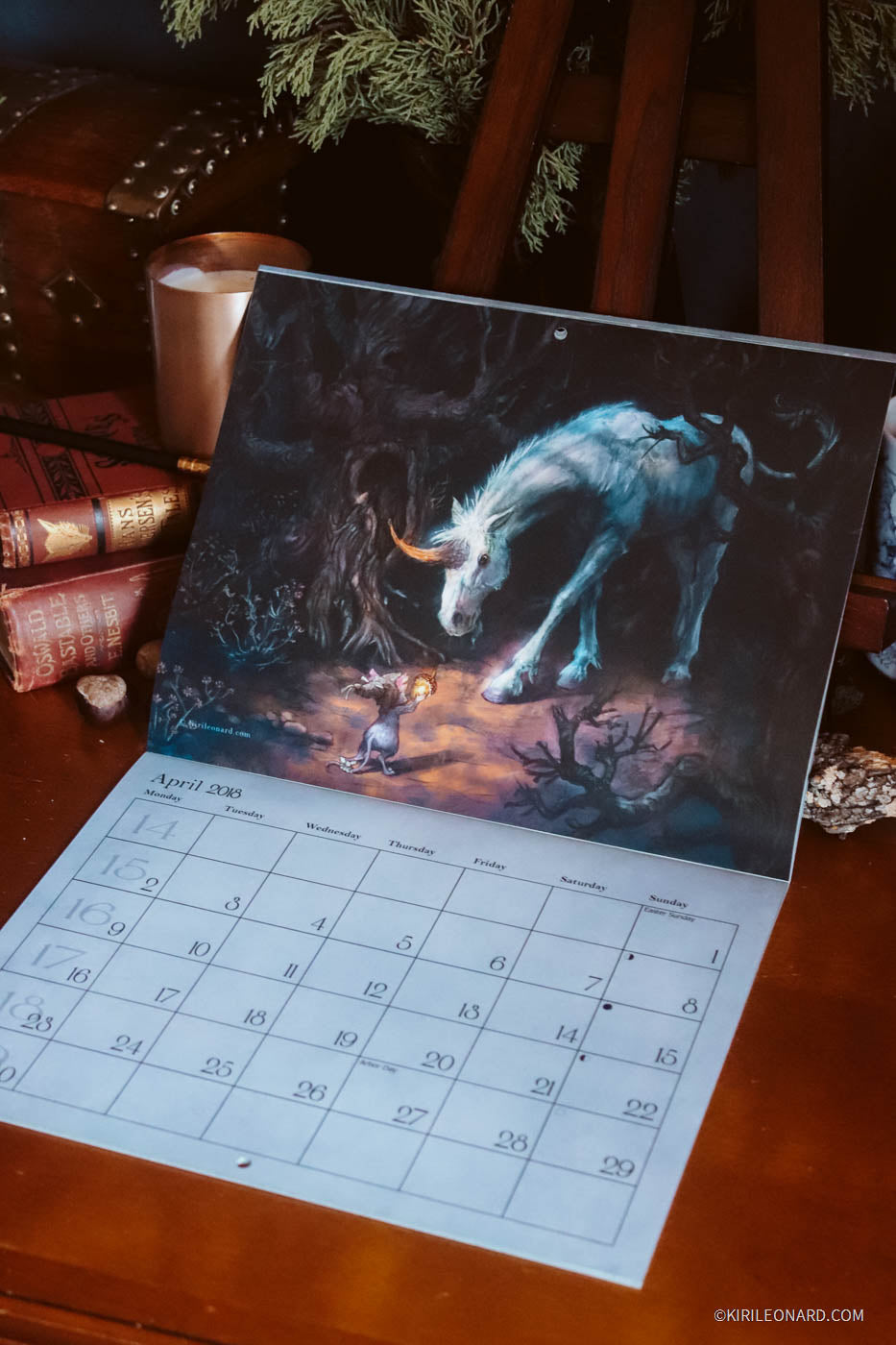 A tabletop calendar spread open on a wooden surface, showing a fantasy illustration of a glowing blue-white unicorn and a small figure in a dark scene. The calendar page below includes the April 2020 grid and the artist credit Kiril Leonard.