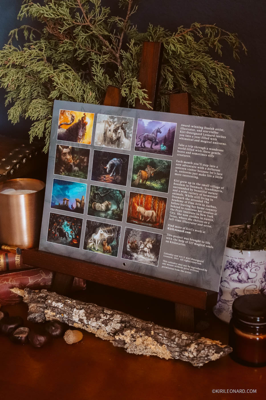 A framed art print or poster displayed on a wooden easel, featuring a grid of twelve colorful animal illustrations and a block of text. The scene is styled with candles, a piece of bark, and greenery against a dark background.