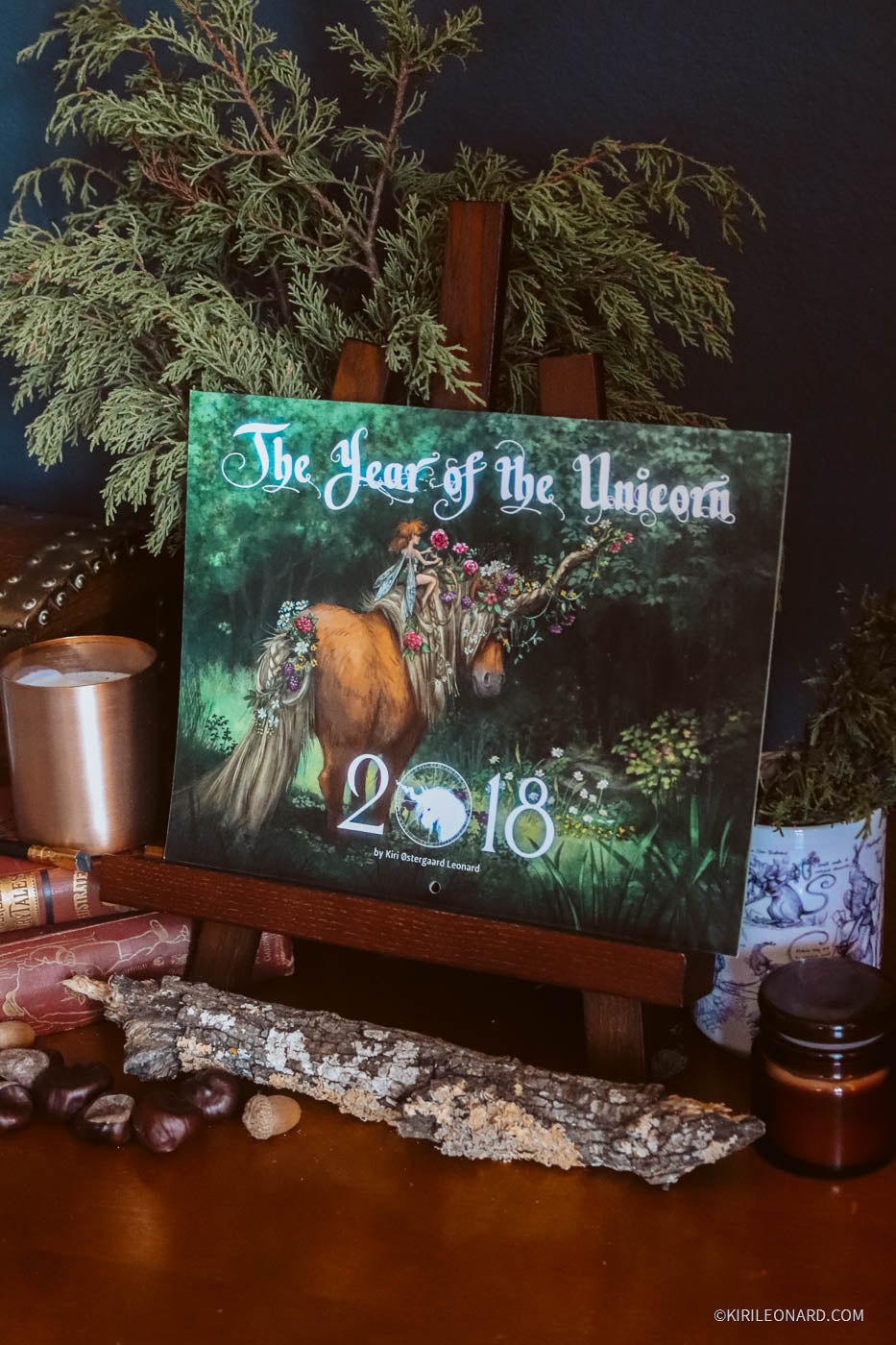 A square art print or canvas titled "The Year of the Unicorn" displayed on a wooden easel. The artwork depicts a unicorn in a forest with the year 2018 overlaid.