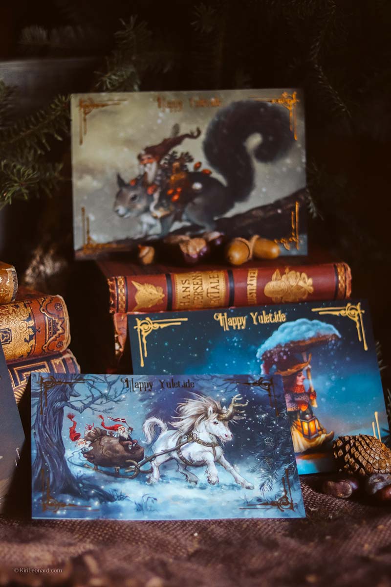 A set of Christmas-themed art prints styled on a rustic dark wooden surface with vintage books, pinecones, and evergreen branches. The prints feature whimsical illustrations: a gray squirrel riding a sleigh with Santa, a white unicorn pulling a sleigh in the snow, and a cozy gingerbread house under a glowing Christmas tree.