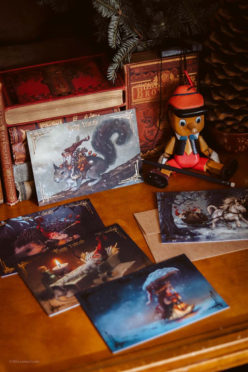 A styled tabletop display featuring multiple fantasy art postcards with illustrated scenes, arranged on a wooden surface alongside vintage books, a pinecone, and a Pinocchio figure.
