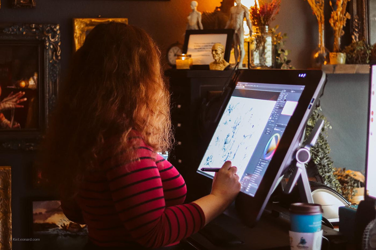 Danish artist Kiri Leonard with long wavy light brown hair, wearing a pink and black striped shirt, is seen from behind as she uses a large digital drawing tablet on a stand in a dimly lit, warm-toned art studio.