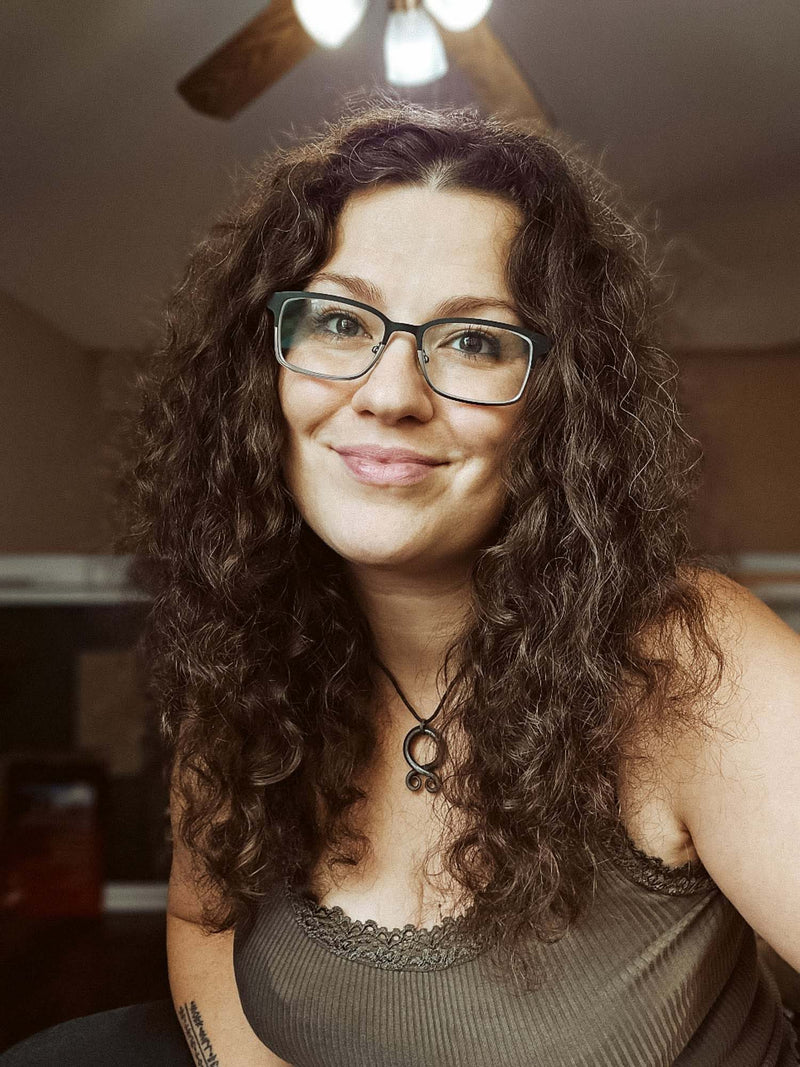 Photo of Danish artist Kiri Leonard with long curly brown hair and black-rimmed glasses, wearing a dark ribbed tank top in an indoor setting.