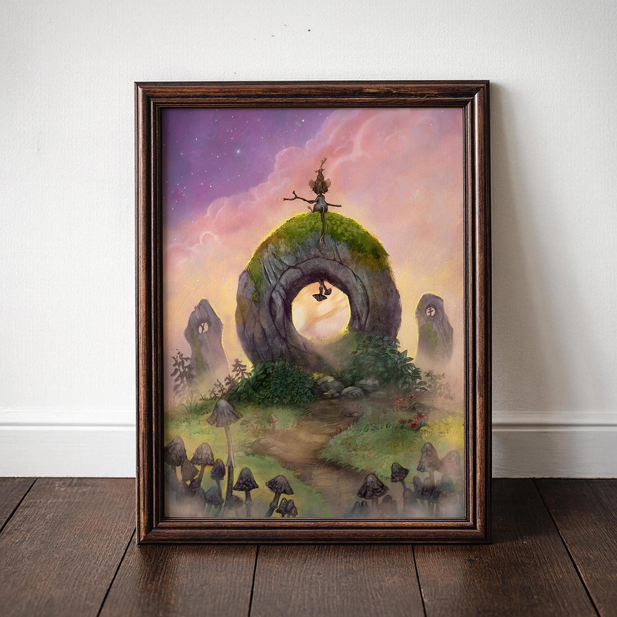 A framed fantasy art print leaning against a white wall on a dark wooden floor. The artwork depicts a whimsical stone ring landscape with a small figure standing on grassy hills under a pink and purple sky, and the frame is a dark brown wood tone.