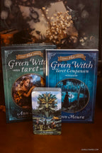 The Green Witch Tarot deck and its companion book displayed on a wooden surface, with the tarot box standing behind the deck and the companion book to the right. A tarot card featuring a tree illustration is placed in front.