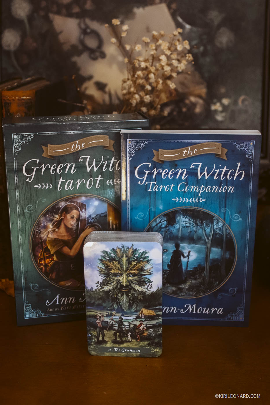The Green Witch Tarot deck and its companion book displayed on a wooden surface, with the tarot box standing behind the deck and the companion book to the right. A tarot card featuring a tree illustration is placed in front.