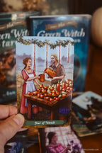 A hand holds a tarot card from The Green Witch deck, showing the 'Four of Wands' illustration. The card features an illustration of a man and woman at a table with apples and candles, with the card's title visible at the bottom.