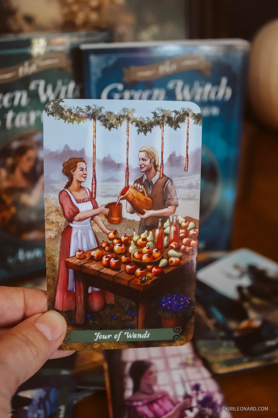 A hand holds a tarot card from The Green Witch deck, showing the 'Four of Wands' illustration. The card features an illustration of a man and woman at a table with apples and candles, with the card's title visible at the bottom.