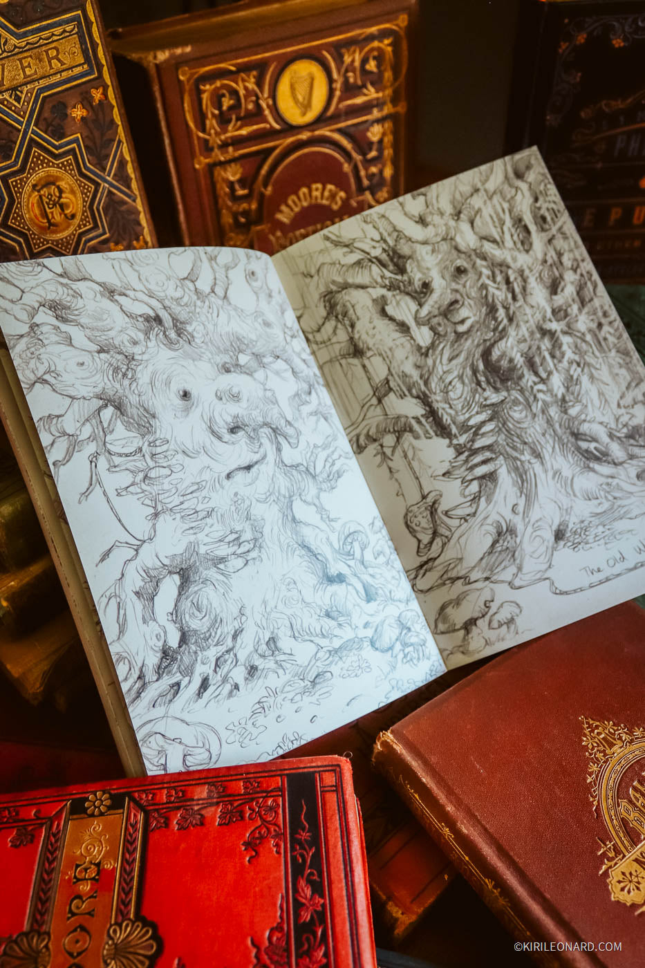 An open sketchbook with detailed black ink drawings of a tree spirit, surrounded by vintage red and gold-bound books.
