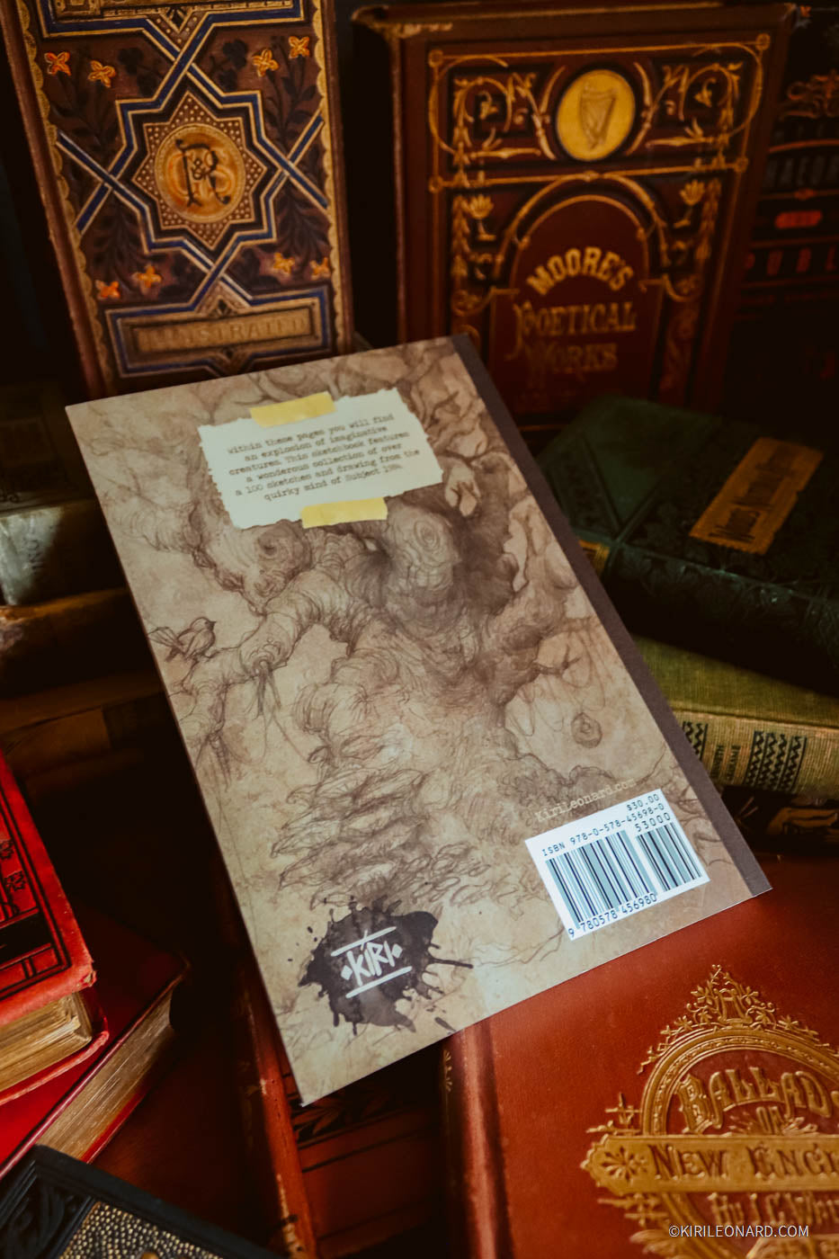 A vintage-style illustrated book or notebook cover shown from the back, resting on top of other old books in a dark, antique-filled setting. The cover has sepia sketch artwork, a small cream label, a barcode sticker, and a black logo near the bottom.