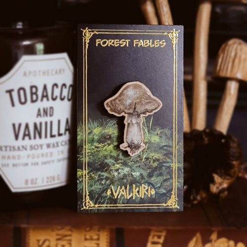 A forest-themed enamel pin shaped like a mushroom, mounted on a decorative black card labeled 'Forest Fables' and 'Valkiri', displayed against a dark rustic background with a wax candle partially visible.
