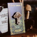 A mushroom-shaped enamel pin displayed on a decorative backing card labeled 'Forest Fables' and 'Valkiria'. The card is photographed on a dark wooden surface next to a bottle label, with blurred mushrooms in the background.