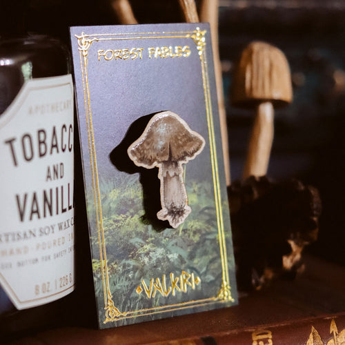 A mushroom-shaped enamel pin displayed on a decorative backing card labeled 'Forest Fables' and 'Valkiria'. The card is photographed on a dark wooden surface next to a bottle label, with blurred mushrooms in the background.