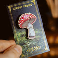 A hand holds a forest-themed enamel pin shaped like a red-and-white toad mushroom on a dark card labeled 'Forest Fables' and 'Valkyr'.