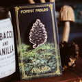 A pinecone-shaped enamel pin with a white border and dark gray/black details, mounted on a decorative backing card. The card reads 'FOREST FABLES' at the top and 'VALKIRI' at the bottom, with a forest illustration.
