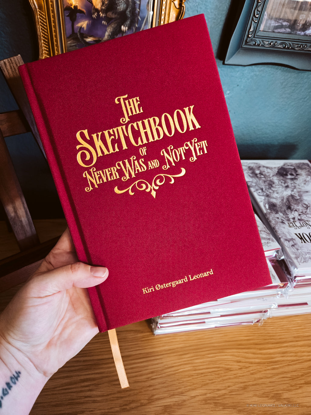 A hand holds a red hardcover sketchbook with gold foil lettering that reads 'The Sketchbook of Never Was and Not Yet' on a wooden table, with framed art and other books in the background.