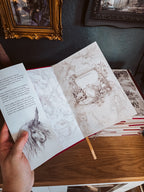 A hand holds an open vintage-style illustrated book or journal on a wooden table, with sketch pages visible and framed wall art in the background.