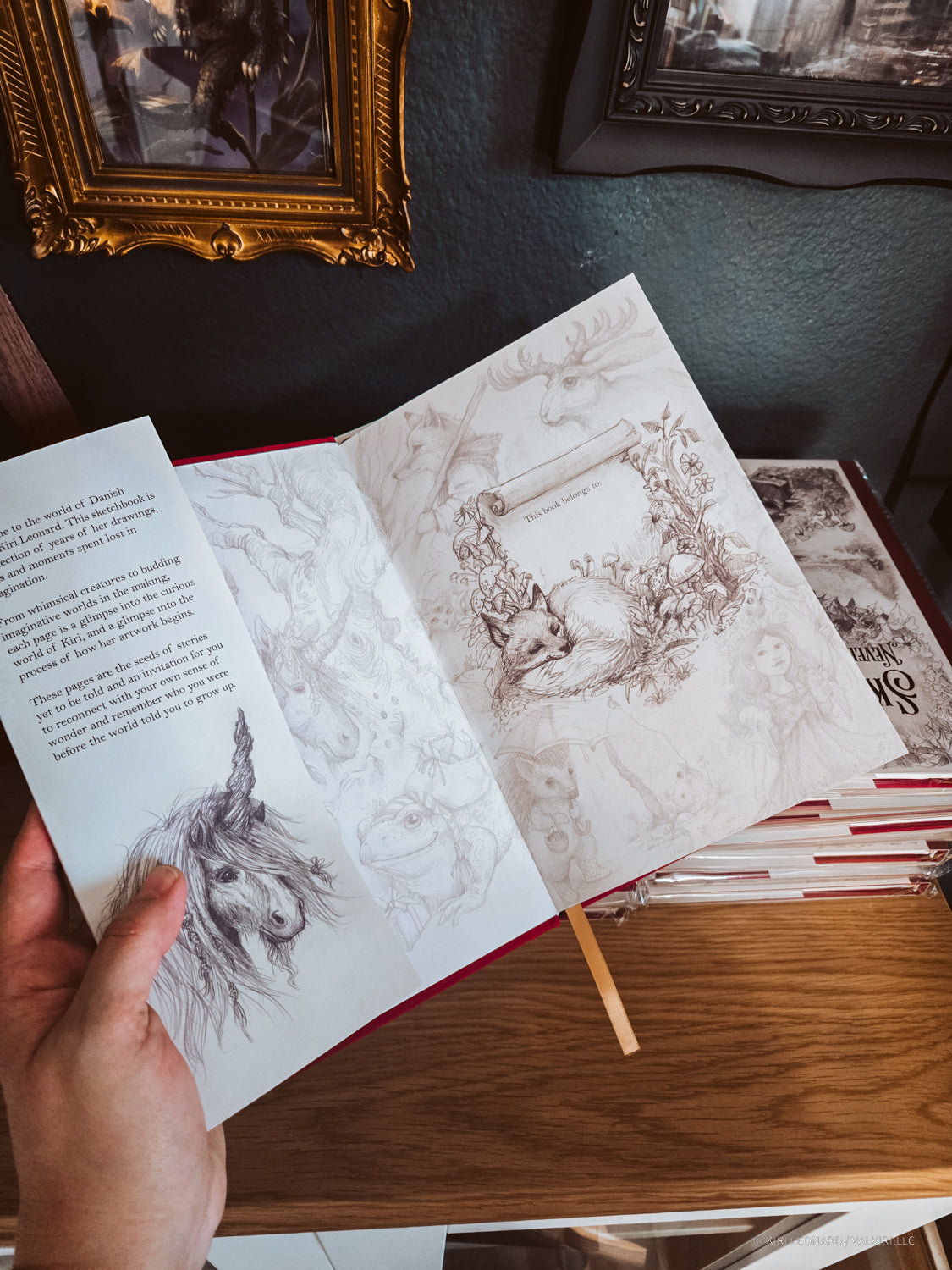 A hand holds an open vintage-style illustrated book or journal on a wooden table, with sketch pages visible and framed wall art in the background.