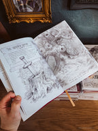 An open illustrated book or journal spread on a wooden table, showing black-and-white fantasy artwork and text including 'The Wooden Sword.' A hand holds the left page, with stacked books and a dark wall in the background.