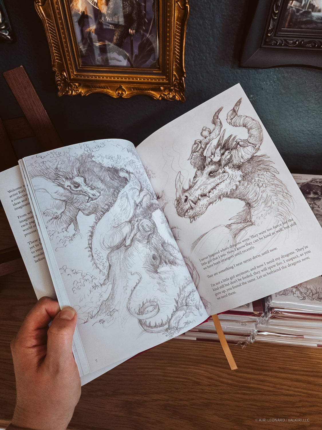 An open sketchbook or art book held by a hand, showing detailed pencil drawings of dragons on the pages, photographed on a wooden table with framed artwork in the background.