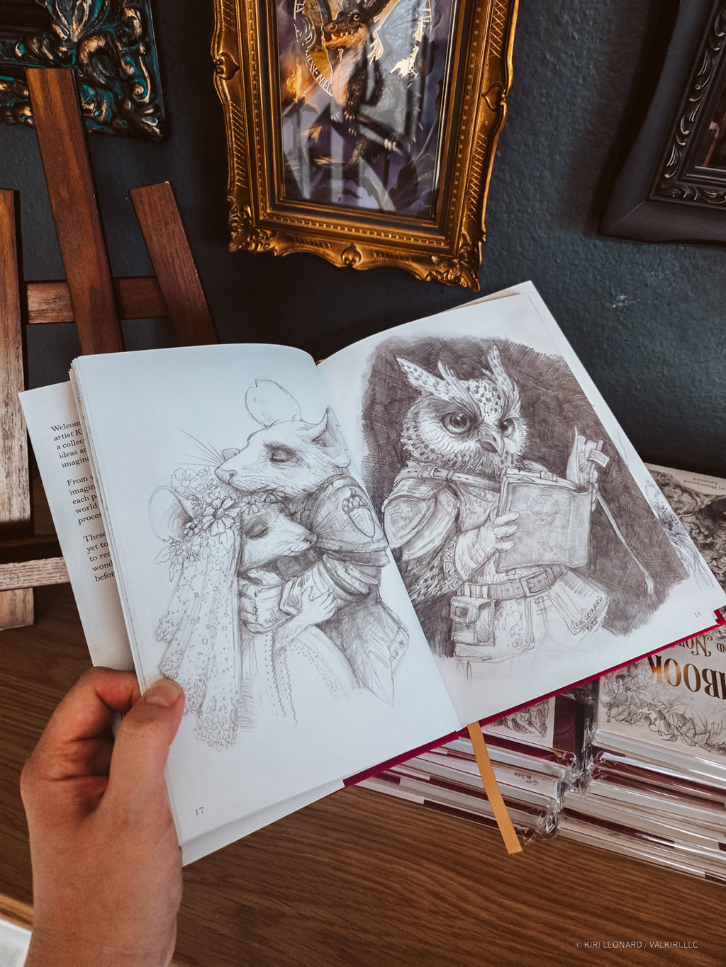 A hand holds an open sketchbook showing detailed pencil drawings of fantasy creatures, with framed artwork and books in the background.