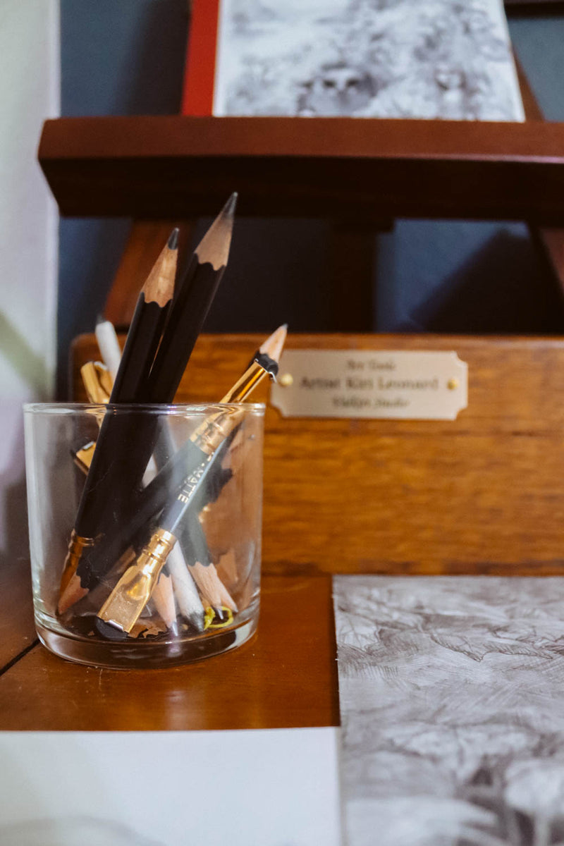 A clear glass cup holding several black and wood-colored pencils, placed on a wooden surface in a real indoor setting with a plaque and artwork visible in the background.