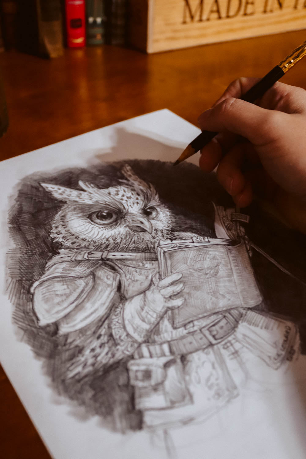 A hand holding a pencil sketches a detailed black and white illustration of an owl wearing medieval armor and holding a book, drawn on a sheet of paper on a wooden desk.