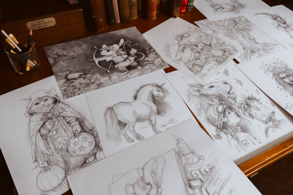 A tabletop display of multiple hand-drawn pencil sketches on white paper, including a cat in a cape, a unicorn, and fantasy characters, arranged on a wooden desk with books and a pencil cup in the background.
